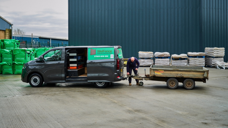 A man attaches a trailer to a gray van labeled "WelFence" in an industrial lot. Pallets and green sacks are stacked nearby. Overcast sky sets a busy tone.