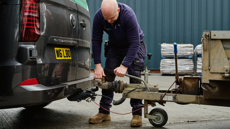 A man connects a utility trailer to a van in an industrial setting. He is focused, wearing work clothes, with stacked materials in the background.