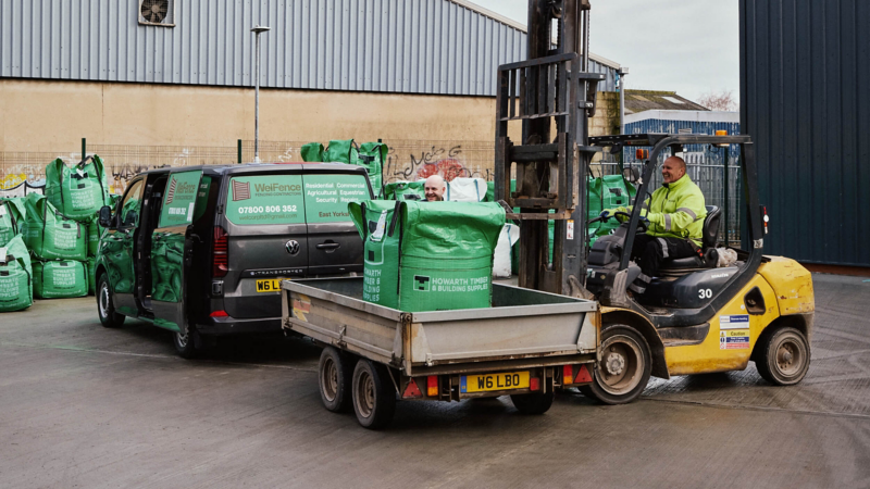 A worker operates a forklift, loading a green bag onto a trailer attached to a van in an industrial yard. Green bags are stacked in the background.