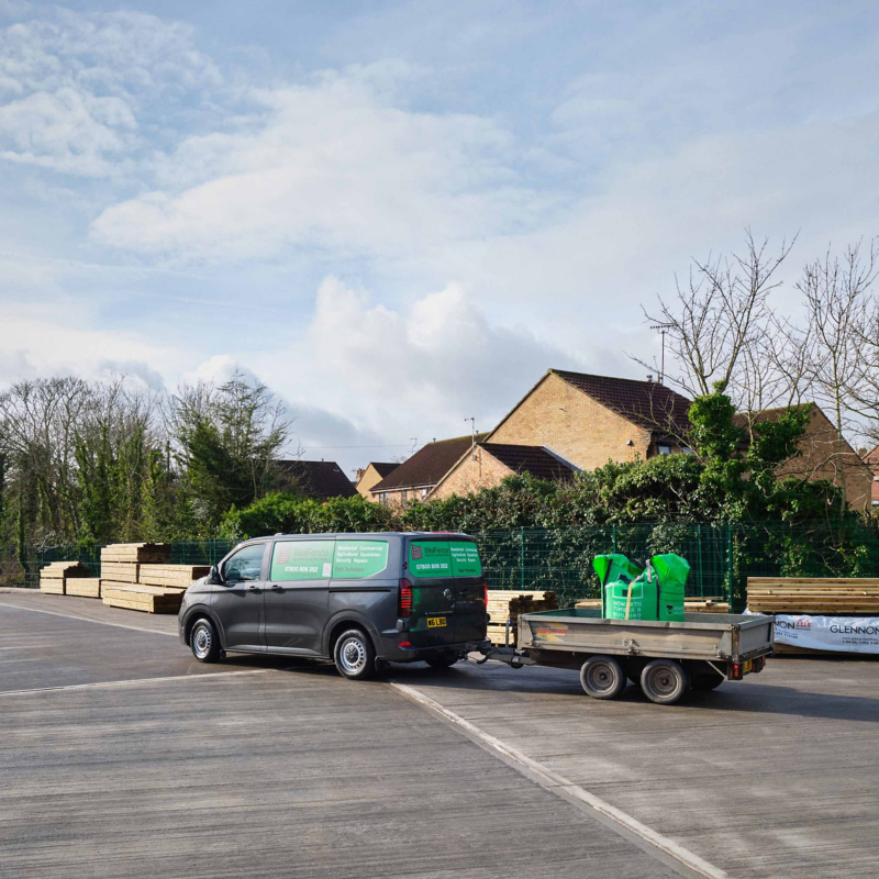 A van with a trailer loaded with green equipment stands on a wide concrete lot. Stacks of lumber are beside it, and houses and trees are in the background.