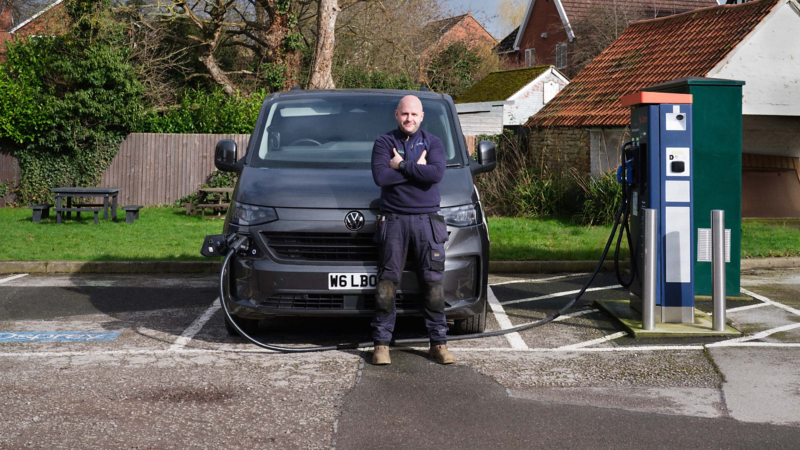 A man stands confidently with crossed arms in front of a black Volkswagen van that's charging at a station, set in a rural area with houses and trees.