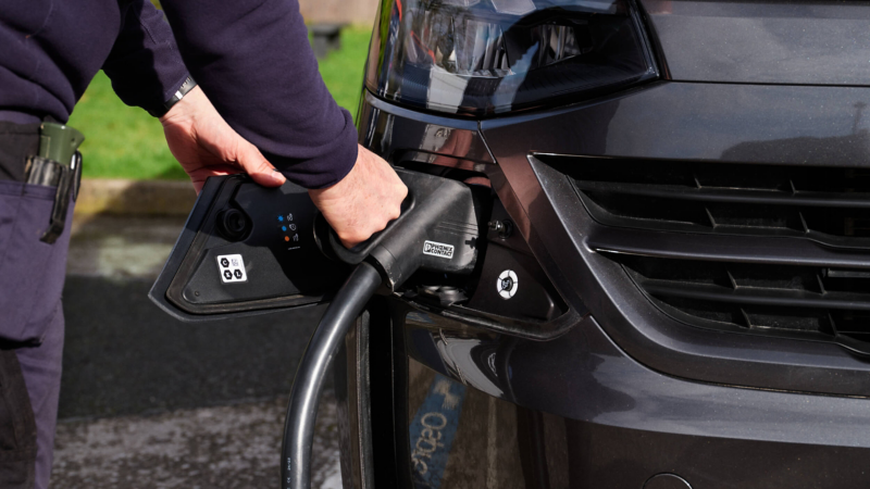A person plugs a charging cable into a black electric vehicle, focusing on the hand and car's front. The scene conveys modernity and eco-friendliness.