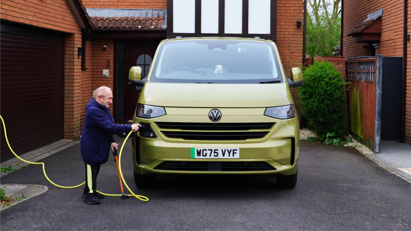 A man holding a charging plug into a front facing green e-Transporter Kombi van