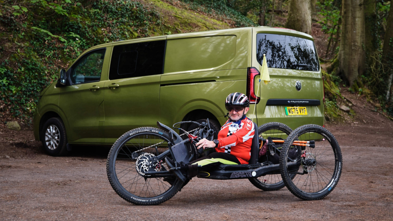 A man sitting in a handcycle in a rural location with a green VW e-Transporter Kombi in the background