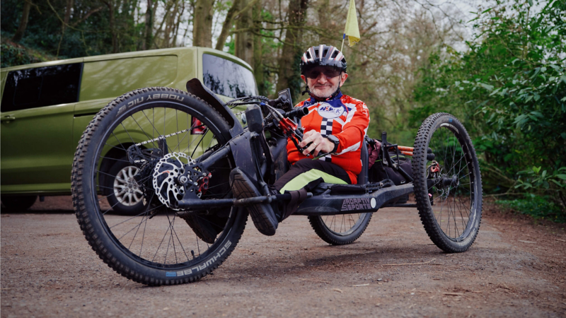 A man sitting in and holding the steering handles of a handcycle with a VW e-Transporter Kombi in the background