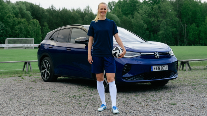 Amanda Ilestedt poserar med en fotboll framför en blå VW ID.4 parkerad vid en fotbollsplan