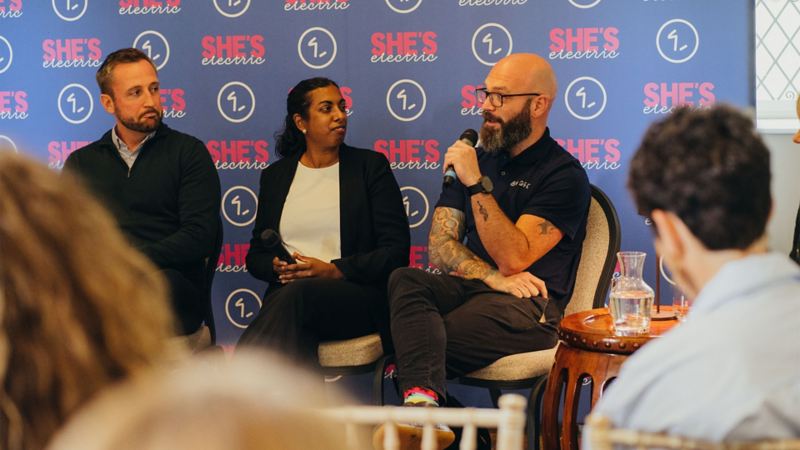 A panel of people on a stage speaking in front of a 'She's Electric' banner