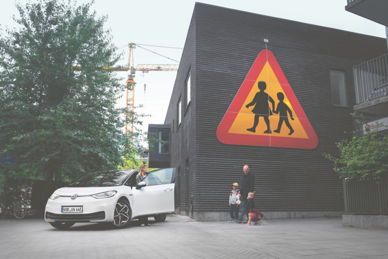 En vit Volkswagen ID.3 parkerad på en innergård, med en familj och en stor målning av ett övergångsställe på en husvägg i bakgrunden