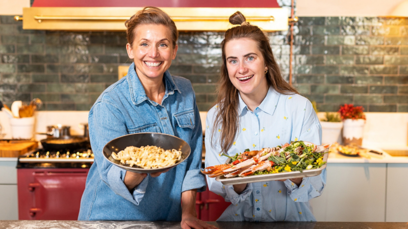 Ebba andersson och Tina Nordström står i ett kök och visar upp maten de lagat