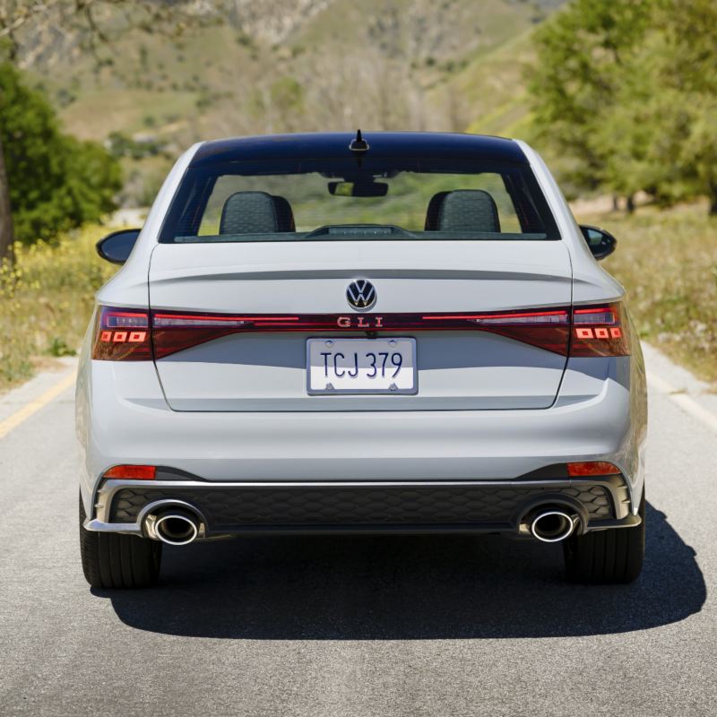 Rear view of a white 2025 Jetta GLI parked on a scenic road with grassy hills in the background.