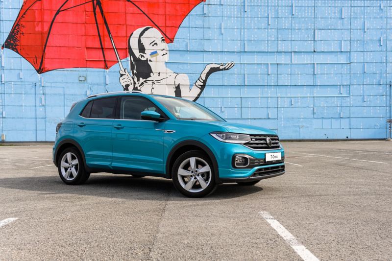 VW T-Cross R-Line in Turquoise in paved car park at coast with beach, view from front right with headlights and front wheel, woman leaning against the vehicle