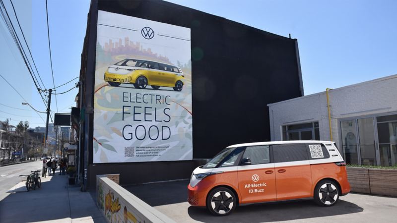 A billboard advertisement for Volkswagen featuring a yellow ID.Buzz with the text ‘ELECTRIC FEELS GOOD' with an orange ID.Buzz parked below the billboard.