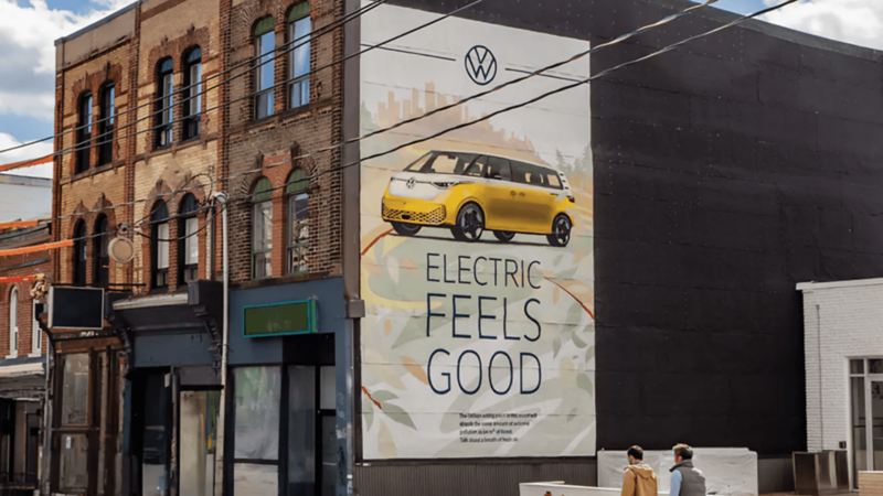 A yellow Volkswagen ID. Buzz is depicted on a mural on a brick building, with the tagline "ELECTRIC FEELS GOOD." Two people walk by.