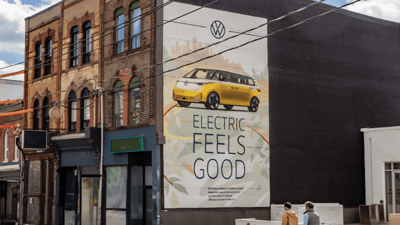 Une fresque sur un bâtiment en briques présente un Volkswagen ID. Buzz jaune avec le slogan "ELECTRIC FEELS GOOD." Deux personnes passent.