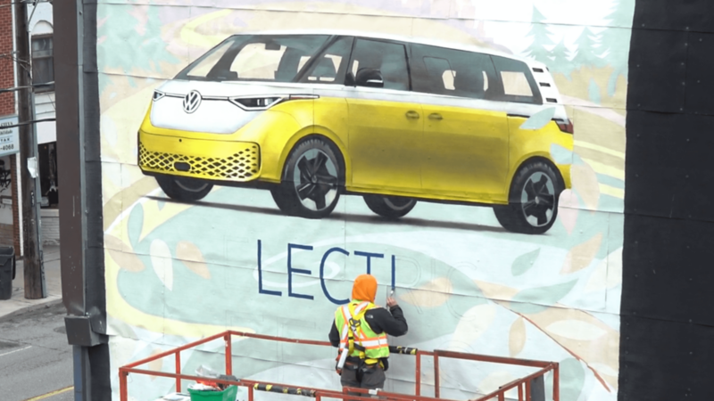 A worker on a lift paints a mural of a yellow Volkswagen ID. Buzz on the side of a building.