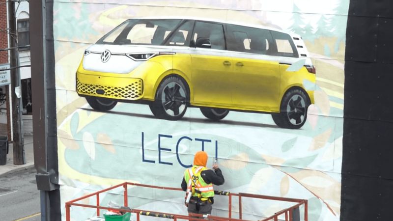 A worker on a lift paints a mural of a yellow Volkswagen ID. Buzz on the side of a building.