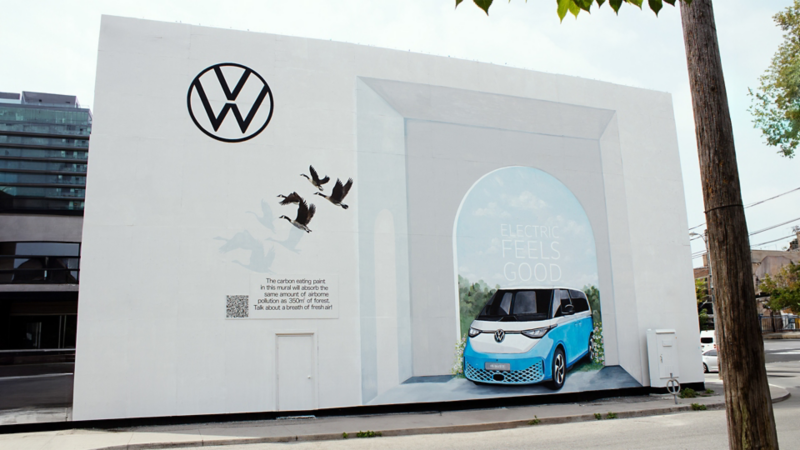 Une grande fresque publicitaire représentant le Volkswagen ID. Buzz traversant une arche blanche, avec le logo VW et quatre oies volant en dessous du logo.