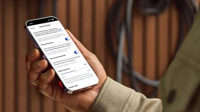 A hand holding a smartphone displaying an EV app's "Battery Settings" screen with energy management and charging options.