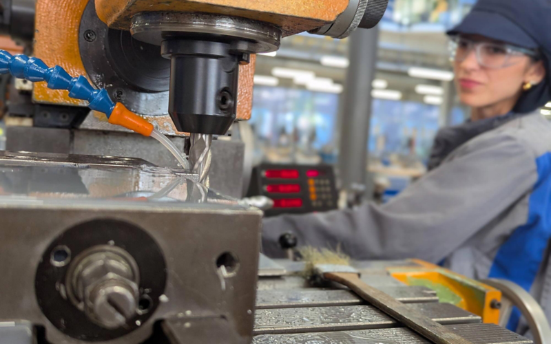 Nahaufnahme einer Maschine zum Fräsen mit einer unscharf zu erkennenden Person in Volkswagen Schutzkleidung, die ein Metallstück bearbeitet 