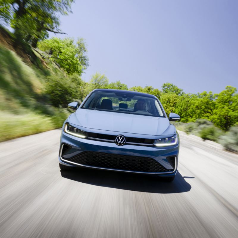 Front view of the blue 2025 Volkswagen Jetta driving on a scenic road with greenery on both sides.