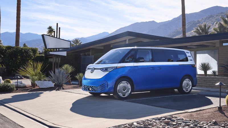 Un VW ID.Buzz bleu étoilé avec une section supérieure blanche nacrée garé devant une maison avec des montagnes en arrière-plan.