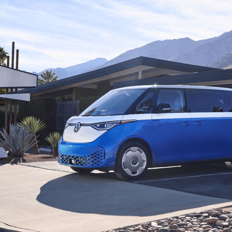 Un VW ID.Buzz bleu étoilé avec une section supérieure blanche nacrée garé devant une maison avec des montagnes en arrière-plan.