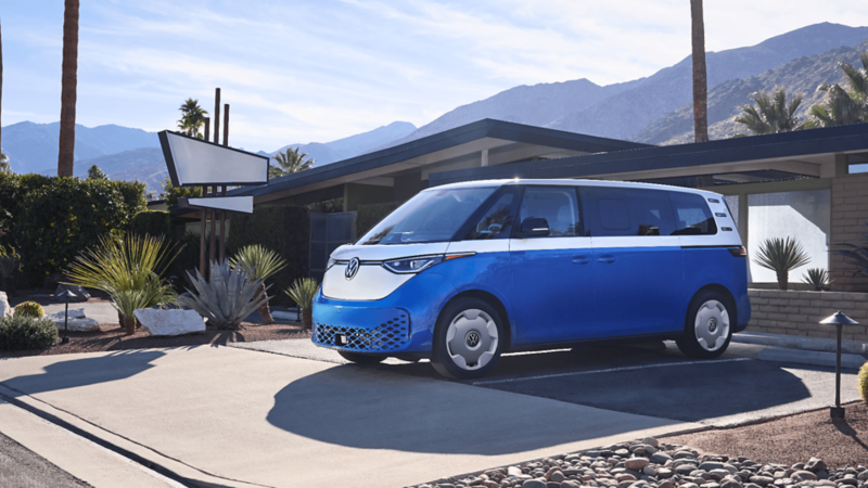 A starlight blue VW ID.Buzz with candy white upper section parked in front of a house with mountains in the background.