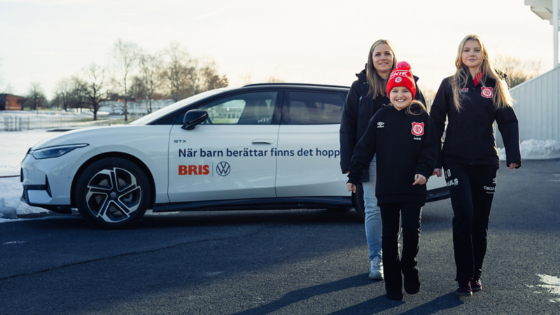 En vit Volkswagen bil med texten "När barn berättar finns det hopp", fotbollstränare Cilla och fotbollsspelarna Astrid och Saga går framför bilen