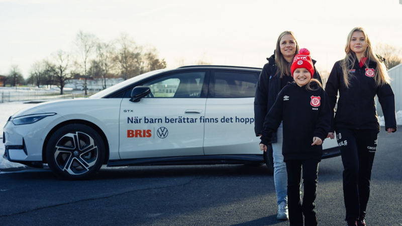 En vit Volkswagen bil med texten "När barn berättar finns det hopp", fotbollstränare Cilla och fotbollsspelarna Astrid och Saga går framför bilen