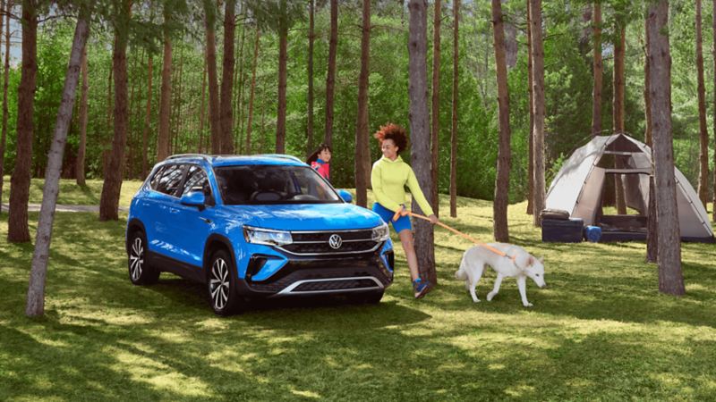 Blue Taos in the forest parked beside a tent. Two females are beside the vehicle, one walking a dog. 