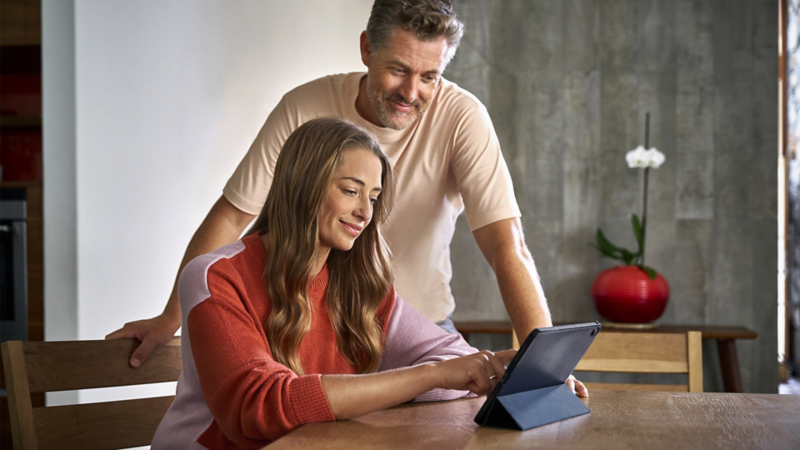 A woman is using a tablet at the table, while a man stands behind and watches.
