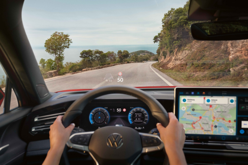 Blick aus dem Fahrzeuginnenraum eines Fahrzeugs auf die Straße mit Darstellung des digitale Cockpits und Navigationsdisplays, die Fahrinformationen und eine Kartenansicht zeigen, während ein Head‑up‑Display die erlaubte Höchstgeschwindigkeit einblendet