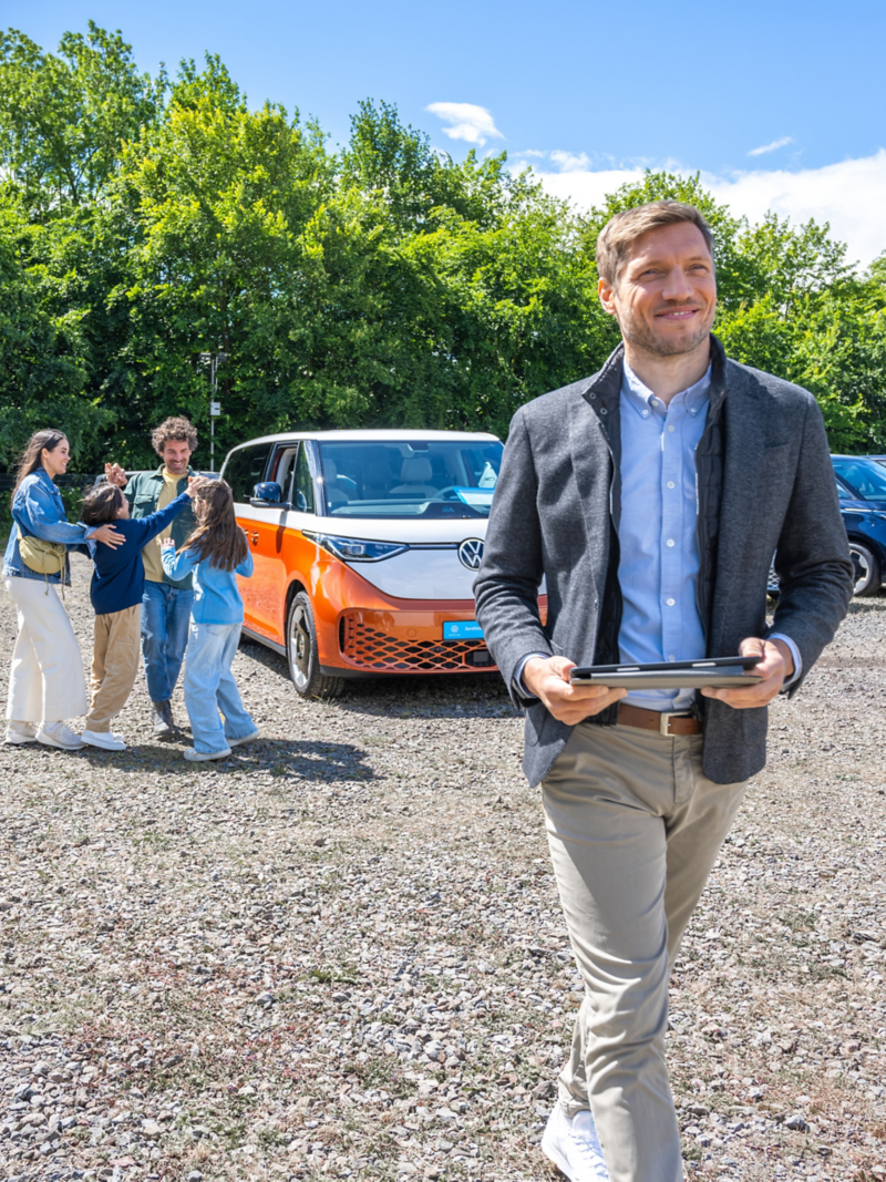 ProfiPartner auf einem Autohof, ein Mann mit Tablet in der Hand im Vordergrund, im Hintergrund eine sich freuende Familie und verschiedene parkende Volkswagen Nutzfahrzeug Modelle.