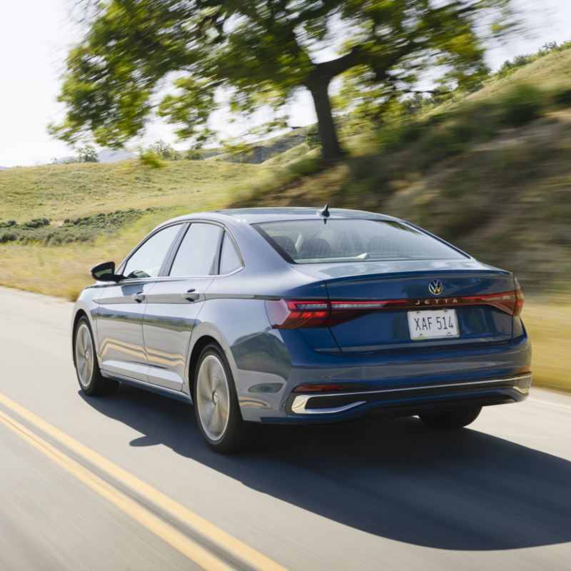 Rear view of the blue Jetta 2025 driving on a scenic road with a large tree and grassy hills in the background.