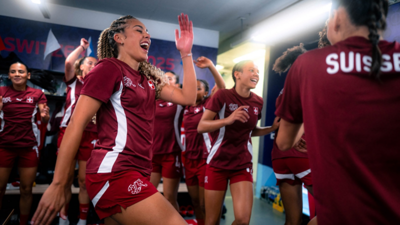Joueuses de l’équipe nationale féminine suisse de football célébrant dans le vestiaire, riant et dansant ensemble en tenues d’entraînement rouges con la croix suisse.