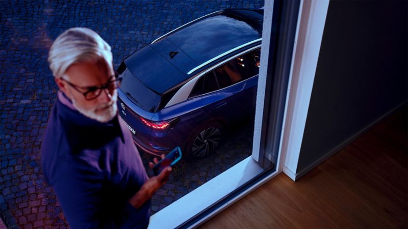 Hombre con un teléfono móvil en la mano junto a la ventana, un VW ID.4 azul al fondo