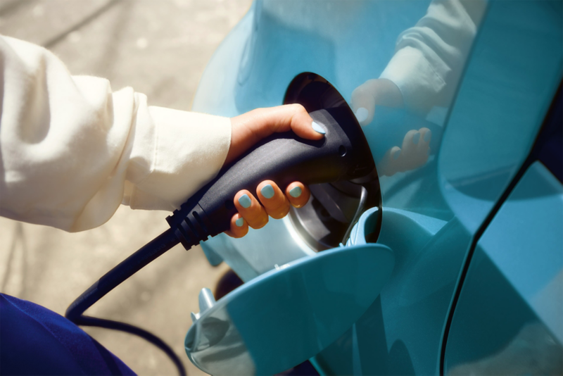  Une main avec les ongles bleus branche un câble de recharge noir dans la prise de charge d'une voiture électrique bleue.