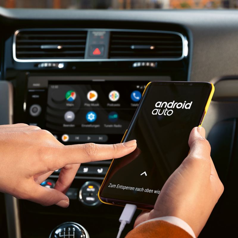 Hand operating connected smartphone with Android Auto logo inside a Volkswagen.