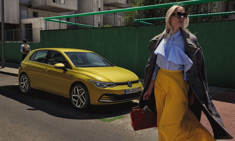 Side view of a yellow VW Golf on a road next to a green wall and a fashionably dressed woman with sunglasses.