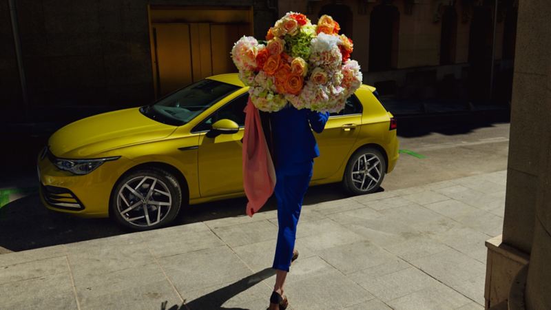 Une femme avec un grand bouquet marche devant une Volkswagen Golf jaune stationnée dans une rue pavée