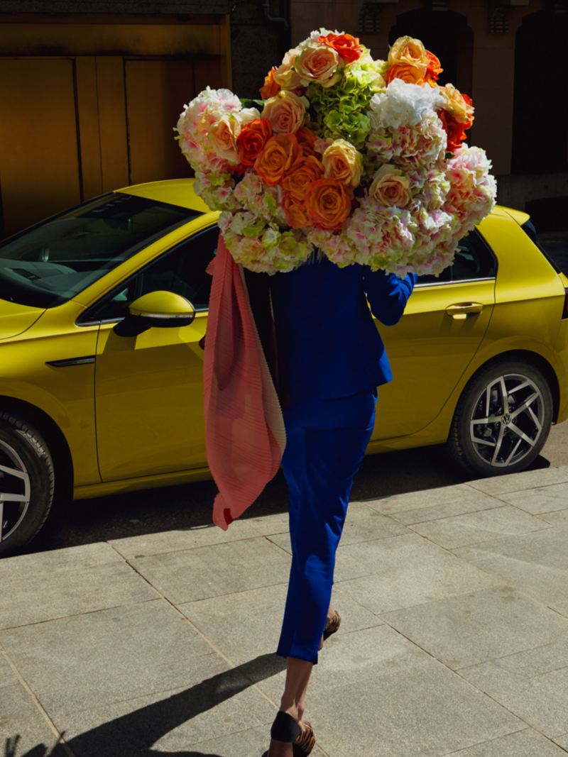 Moteris su glėbiu gėlių eina prie „VW Golf“