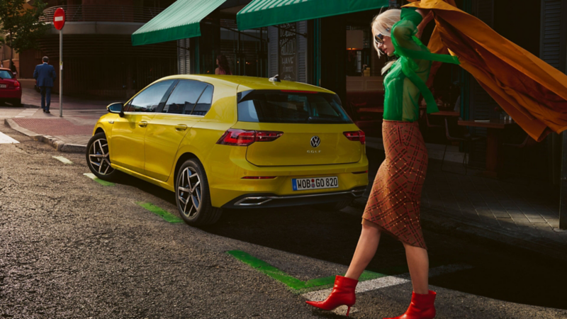 Golf jaune stationnée, vue de 3/4 arrière. Une femme passe derrière avec un voile à la main volant au vent.