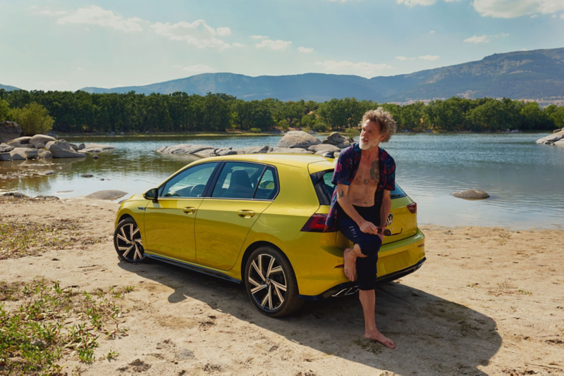 Man at the rear of the VW Golf, in the background a lake and mountains