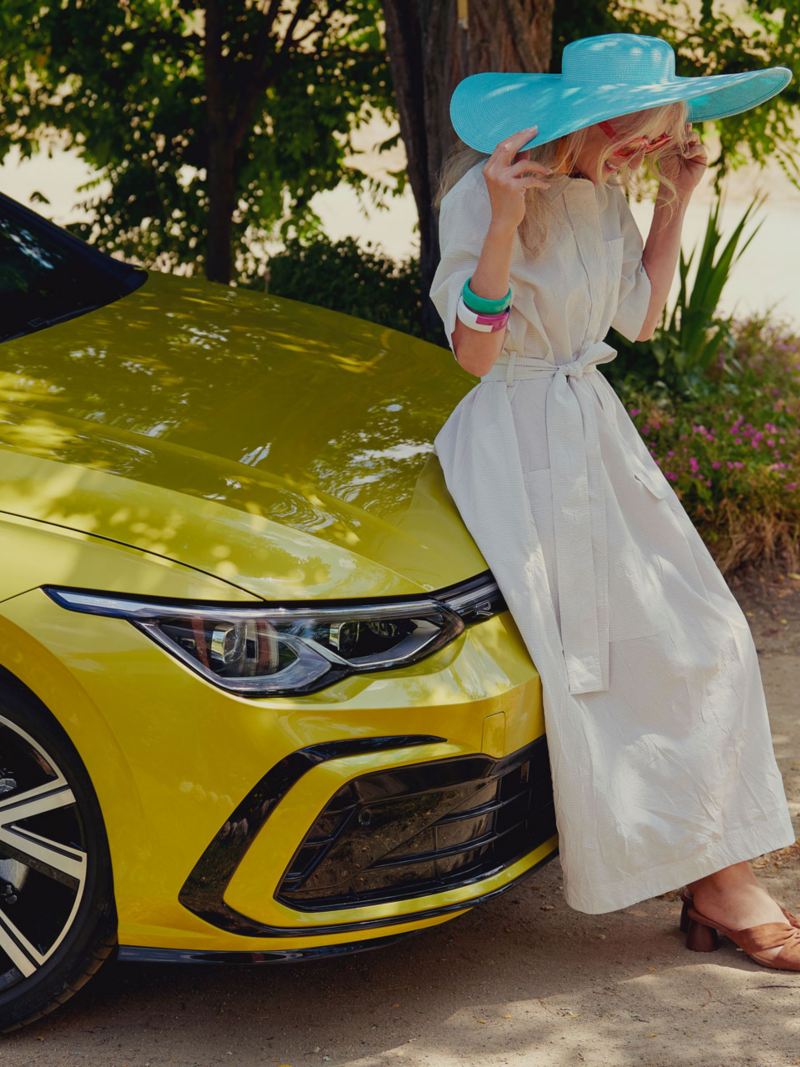  Sieviete baltā kleitā atspiedusies pret jaunā VW Golf motora pārsegu