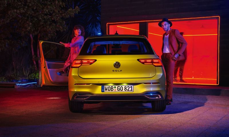 Full-on rear view of a yellow VW Golf with a couple getting out, a warmly lit window in the background.