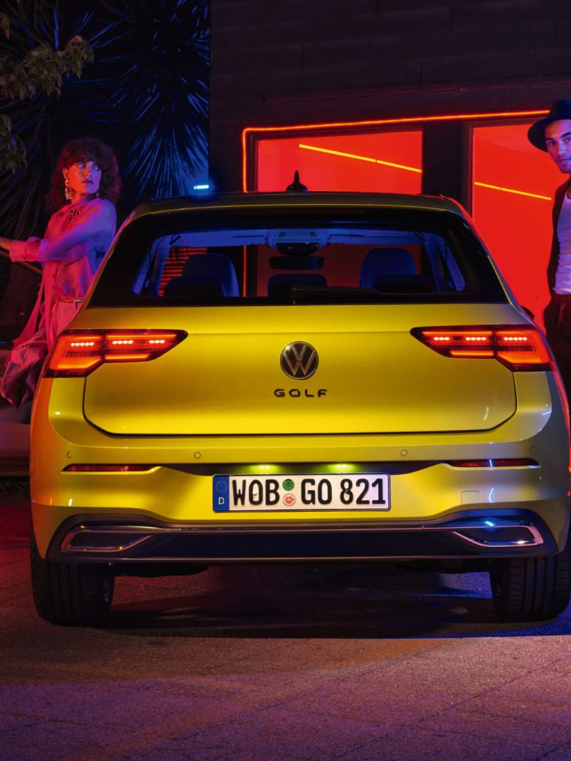 Full-on achteraanzicht van een gele Volkswagen Golf met een stel dat uitstapt, een warm verlicht raam op de achtergrond.