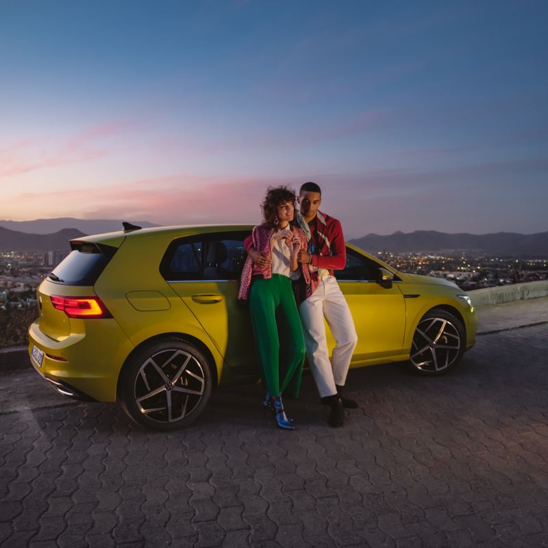 Paar lehnt an VW Golf vor abendlichem Stadtpanorama