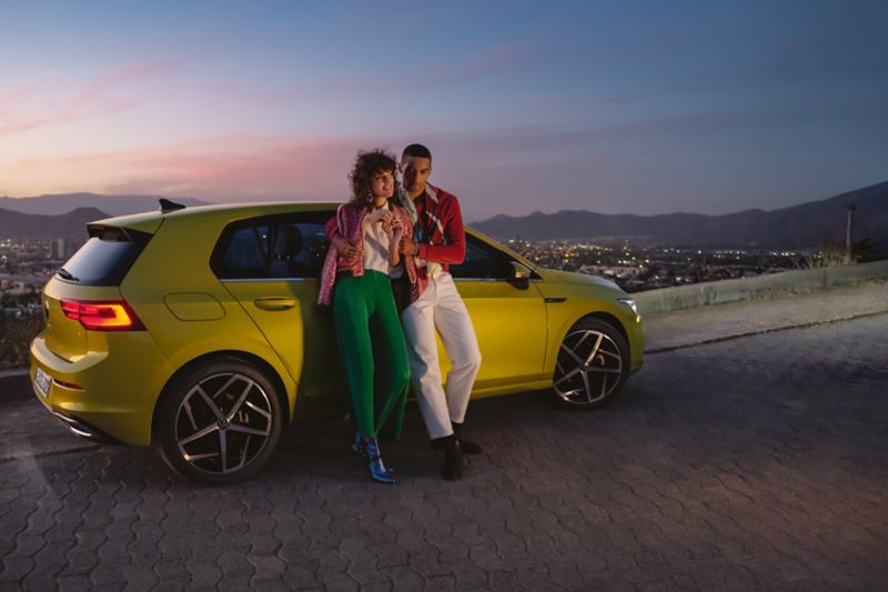 VW Golf jaune sur une montagne dans la lumière du soir avec un couple se serrant dans les bras.