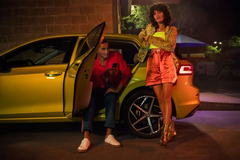 Side view of a yellow VW Golf, a young man exiting the rear door, a young woman standing next to it.