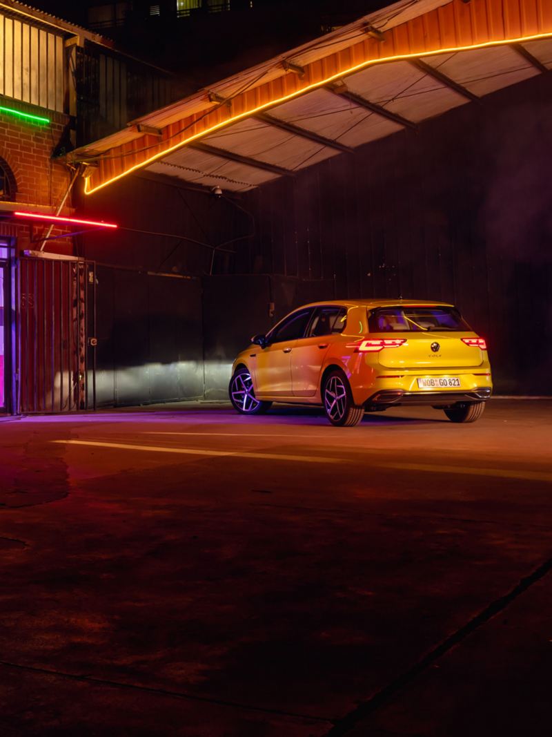 Rear view of a yellow VW Golf at night in a car park, the LED headlights illuminating a building.